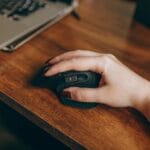 Dłoń z myszką bezprzewodową obok laptopa na drewnianym biurku