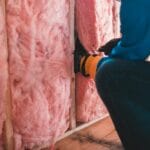 Jak poprawnie ocieplić stary dom od zewnątrz: etapy prac, typowe błędy i wskazówki dla inwestora Pracownik w rękawicach montuje różową wełnę mineralną na ścianie